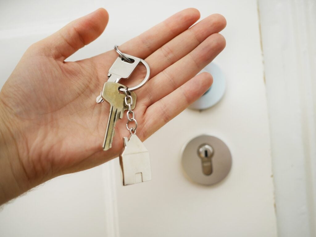 Hand holding house keys with a house-shaped keychain, near a door lock, illustrating the importance of residential locksmith services for home security.